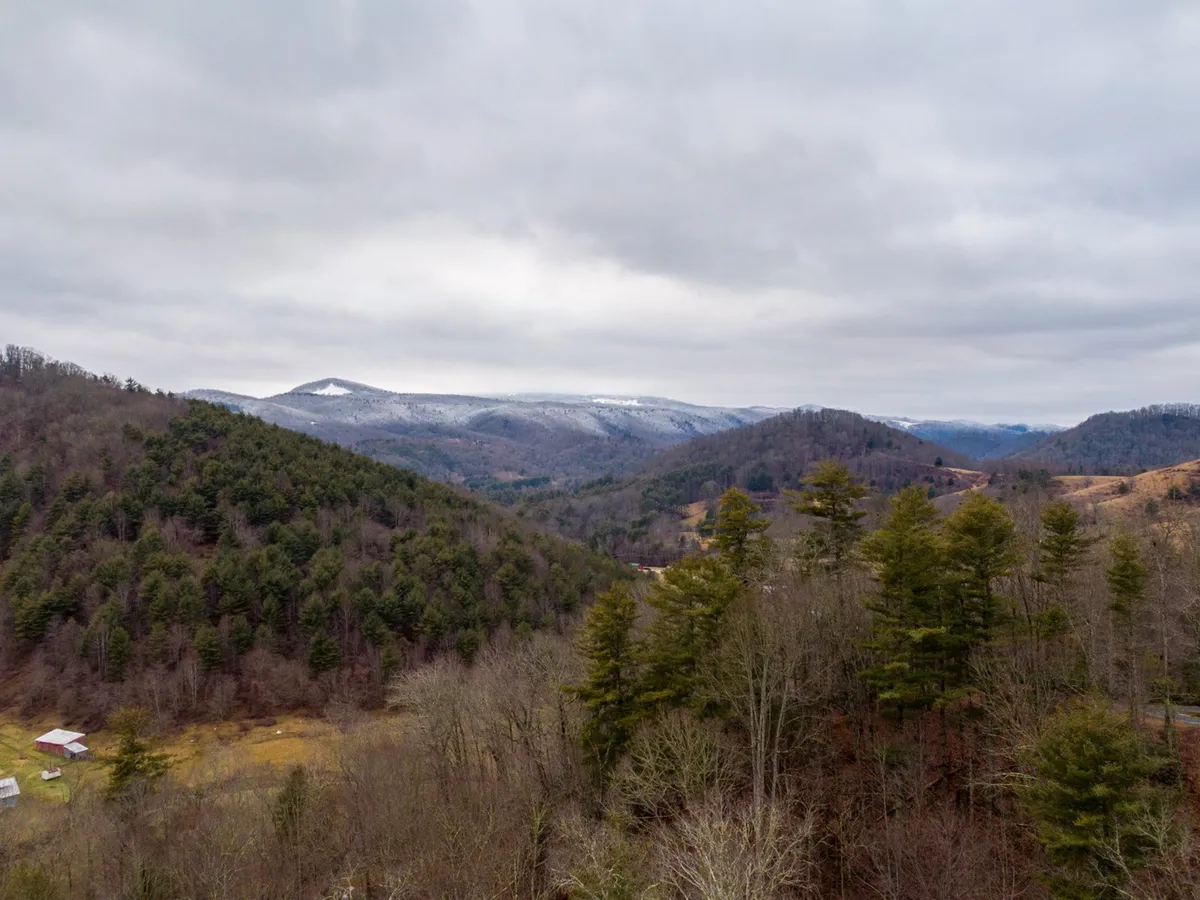 A sweeping scenic view of rolling hills, dense forests, and distant snow-capped mountains, with scattered farmhouses nestled in the landscape. The scene is tranquil and awe-inspiring, highlighting the area’s natural beauty.