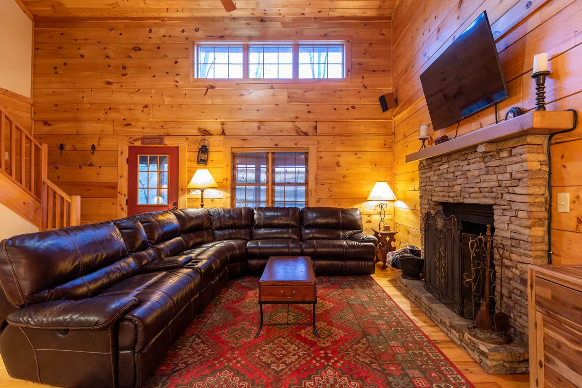 This interior image features a welcoming living room with a leather sectional sofa arranged around a robust stone fireplace. Wooden walls and a soft rug add to the cozy atmosphere, while a Smart TV is visible, making the space ideal for relaxing movie nights or gatherings. The fireplace is central, with firewood and comfortable seating nearby.