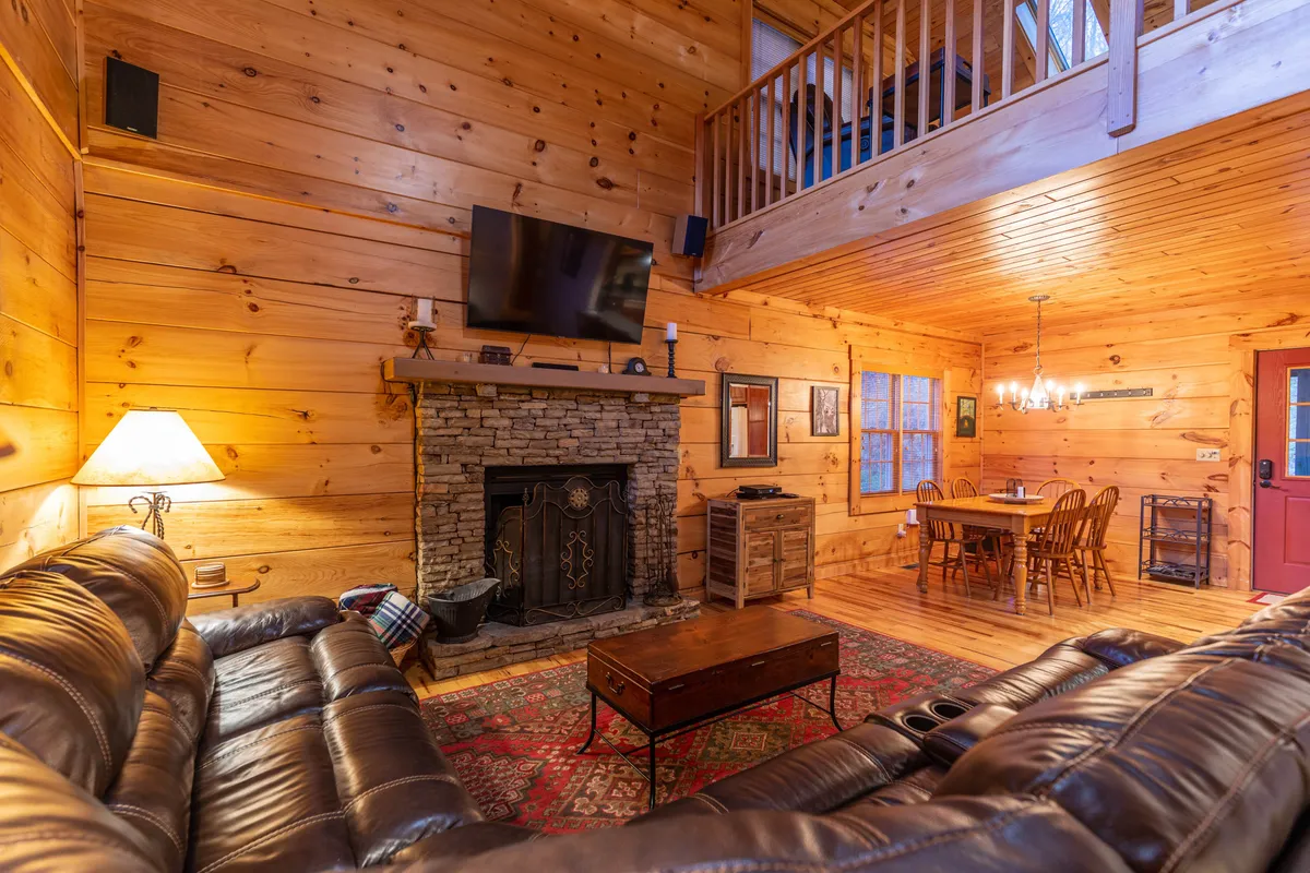 The photo highlights a spacious living and dining area with a stone fireplace, leather sofas, a large dining table, and wooden wall paneling. A TV is positioned for easy viewing from the seating area. The open-plan layout seamlessly connects the living and dining spaces, fostering a sociable environment.