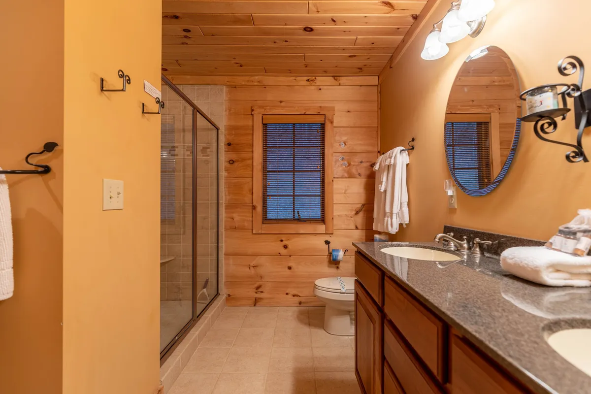 A serene bathroom with a double sink vanity, large mirror, shower area, wooden accents, and a window with blinds. The space feels open and calming, ideal for private moments and daily routines.