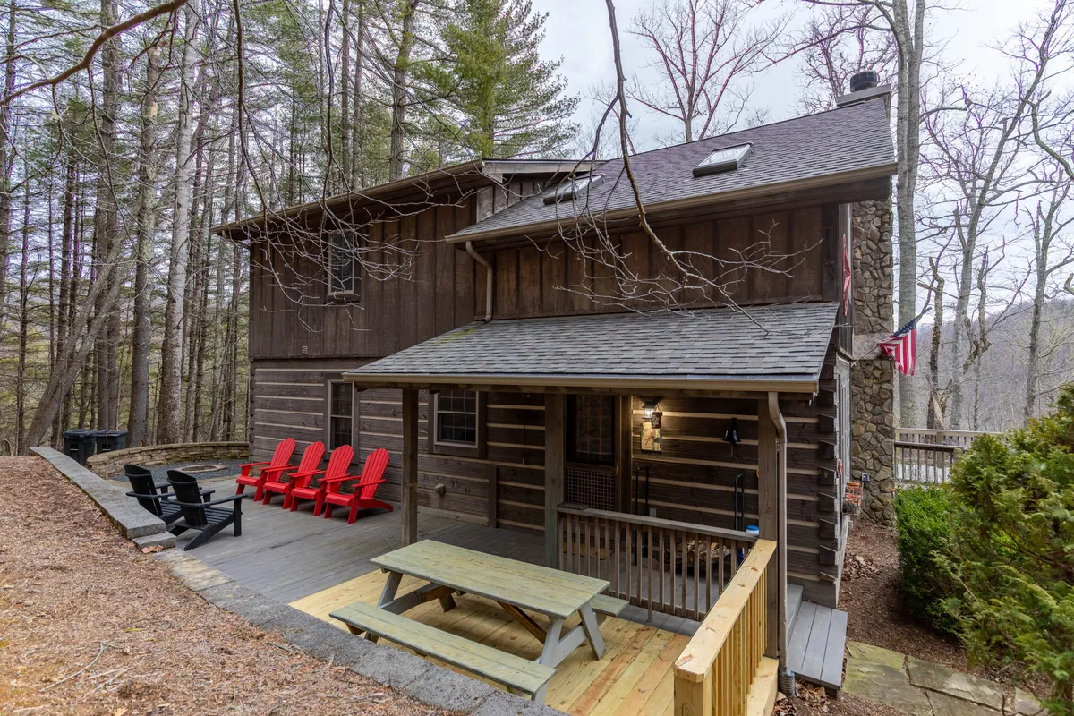 The image shows the cabin exterior with a wooden deck, several red Adirondack chairs, and a picnic table, all set against a backdrop of towering trees. The arrangement invites both group gatherings at the table and intimate conversations by the fire, with plenty of space for children to play safely within the deck’s boundaries.