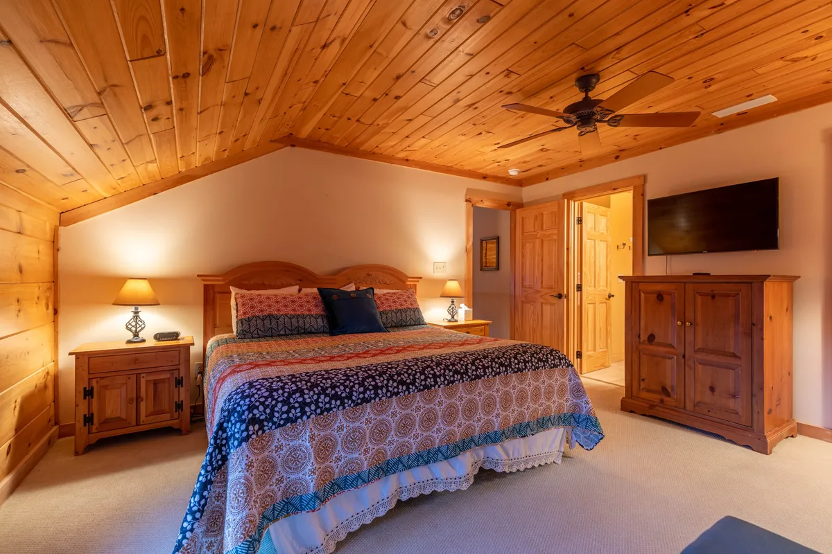 A cozy bedroom with a bed adorned in colorful bedding, complemented by wooden furniture, a nightstand with a lamp, and a Smart TV mounted on the wall. The overall ambiance is warm and comfortable, inviting rest and relaxation.