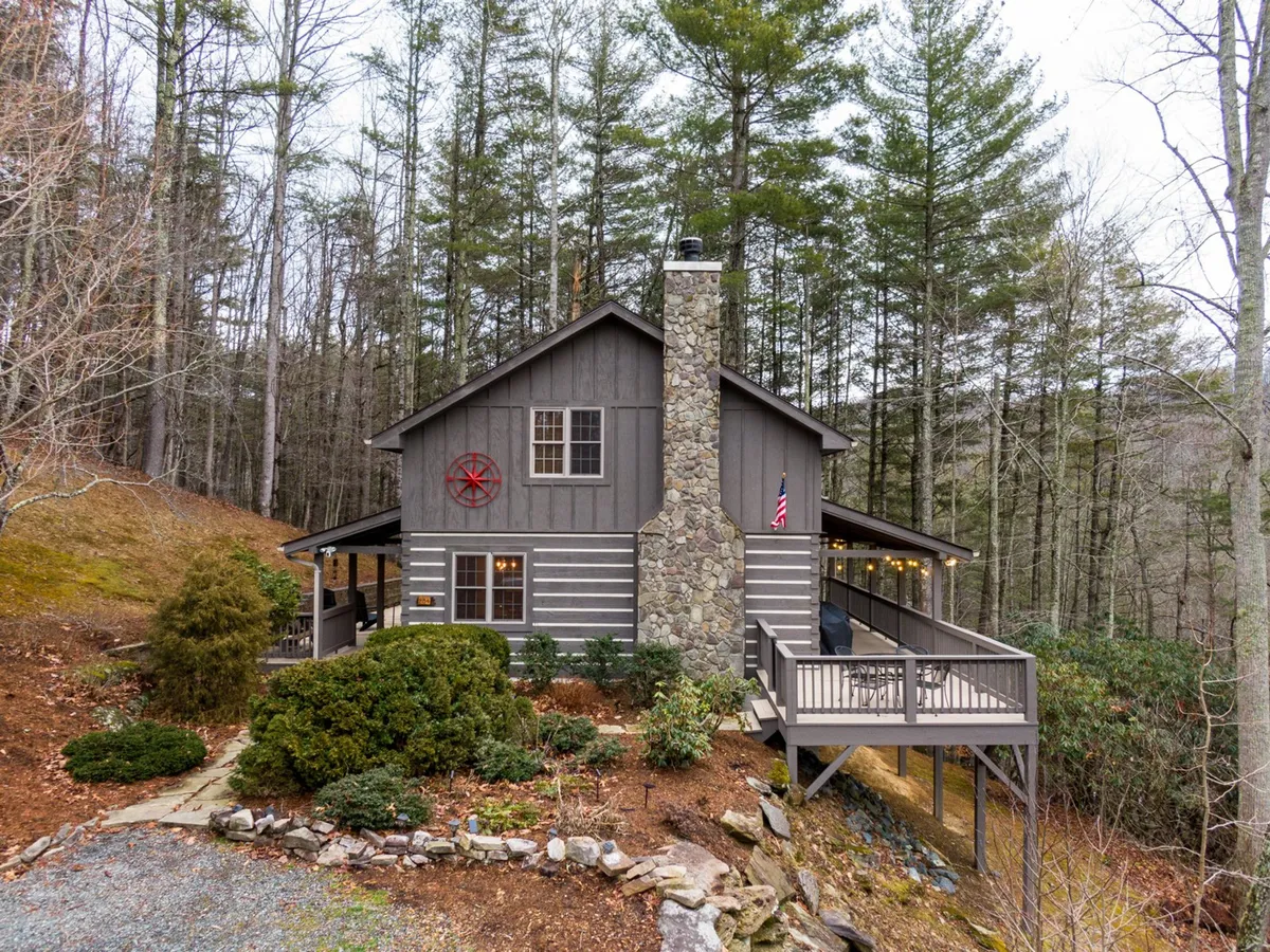 The image shows the exterior of Cozy Cabin, featuring a welcoming stone chimney, a spacious wooden deck adorned with a red star decoration, and beautifully landscaped gardens. The surrounding woods hint at access to nature trails and space for outdoor gatherings.