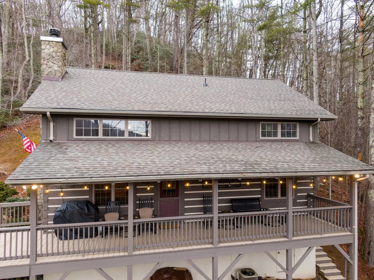 The image shows a welcoming wooden porch with rocking chairs, a stone chimney, an American flag, and a comfortable seating arrangement. The lush greenery and front steps suggest easy access to the outdoors and room for family relaxation.