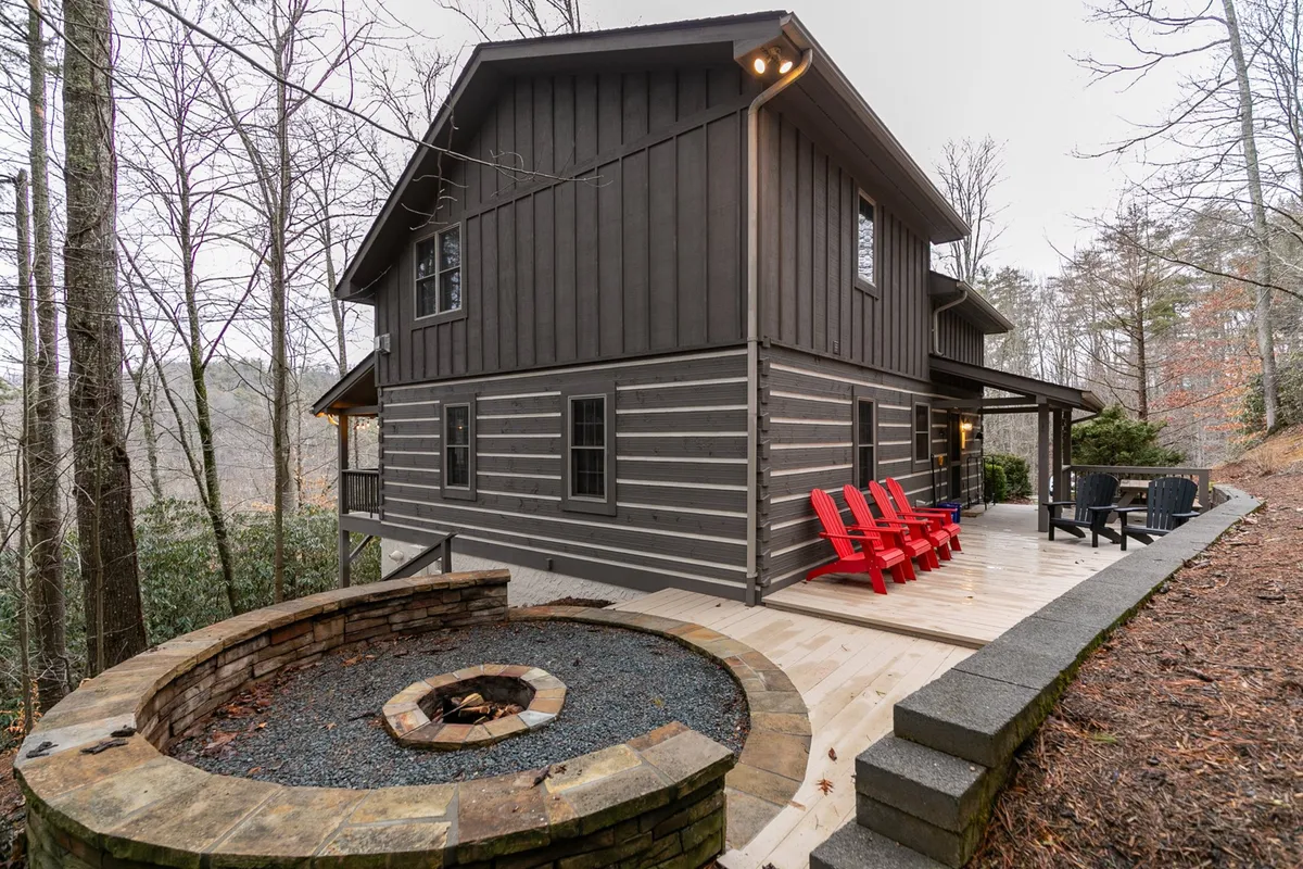 This inviting exterior scene features a well-built fire pit surrounded by comfortable deck chairs, set on the wooded edge of the property. The wooden deck and cabin siding are visible, with mature trees providing privacy and a natural mountain backdrop. The setup is ideal for socializing, outdoor dining, and relaxing by the fire as the sun sets.