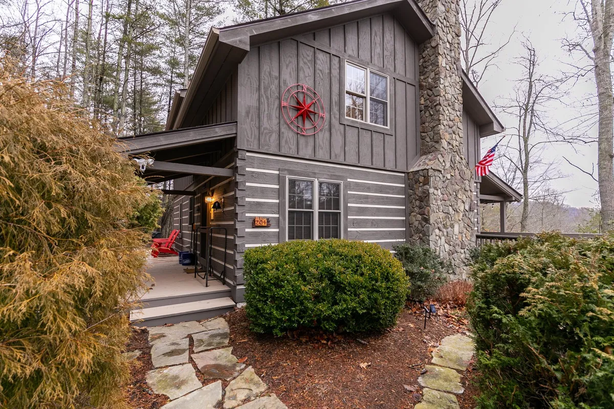 This exterior image captures the front porch area with a stone chimney, red wheel decoration, and rocking chairs set amid lush green shrubs. The setting is tranquil and welcoming, suggesting both the adventure of the wilderness and the comforts of returning home.