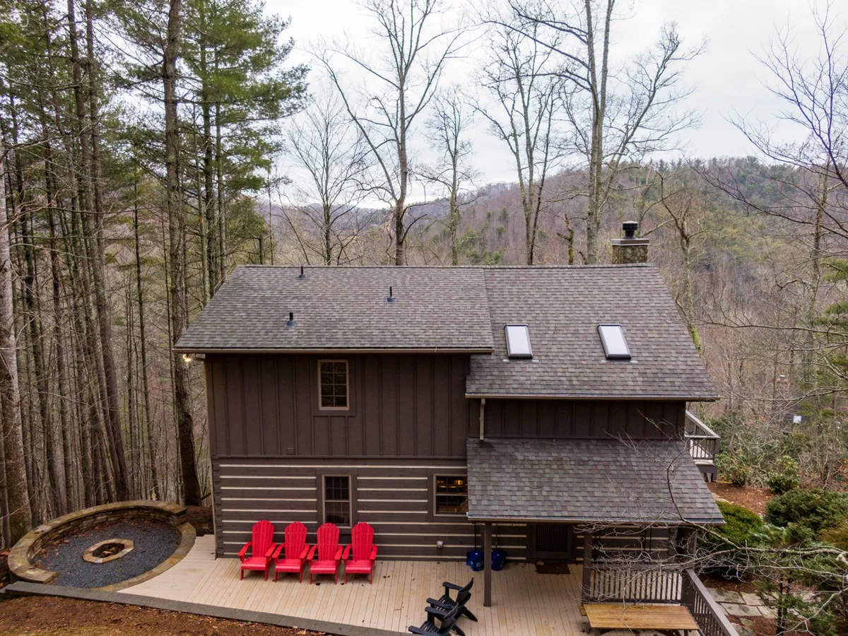 A tranquil view of the wooden deck features red Adirondack chairs clustered around a stone fire pit, surrounded by lush trees. The deck offers a cozy spot for gatherings, with space for both relaxation and conversation. The peaceful setting highlights the privacy and natural beauty of the location.