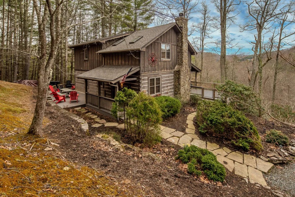 The image displays a charming wooden log cabin nestled among tall trees, with a welcoming stone pathway leading up to a covered deck. Bright red chairs invite guests to relax outdoors, while the tranquil setting hints at both sunset views and the promise of peaceful mornings in nature.