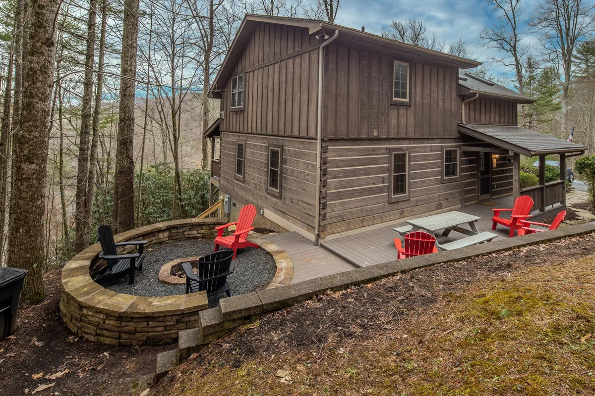 This photo captures a fire pit surrounded by red Adirondack chairs, set against the rustic wooden siding of the house and framed by leafy trees. The scene invites group gatherings, with plenty of seating for friends to share stories or laughter around the fire in a woodland setting.