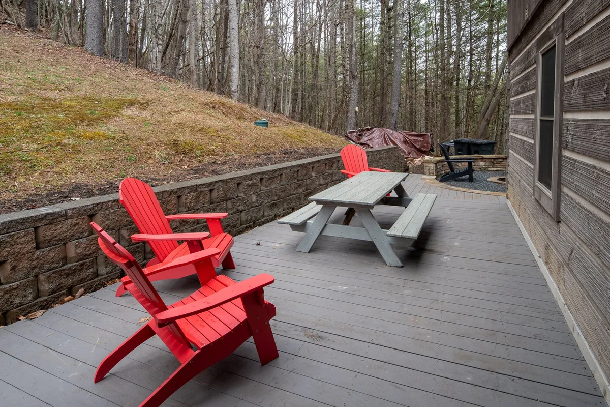 This image features a set of red Adirondack chairs and a gray picnic table on a stone patio, providing a spacious area for outdoor dining and relaxation. The surrounding nature and informal seating create a welcoming space for group meals or casual conversation.